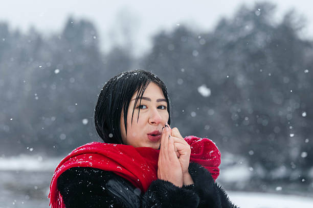 Asian female stands outdoors in snow, warming her hands with a red scarf around her neck, surrounded by a winter landscape and falling snowflakes