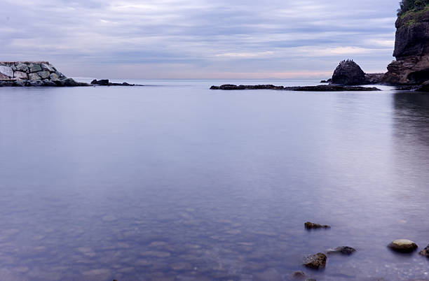 A serene long exposure photograph of a calm rocky coastline under an overcast sky. The slow shutter speed creates a misty, ethereal water effect over submerged pebbles and a stone pier.