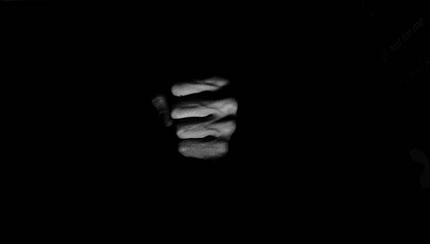 Monochrome, low-key close-up of a human fist emerging from darkness, dramatic spotlight and high contrast with large copy space.