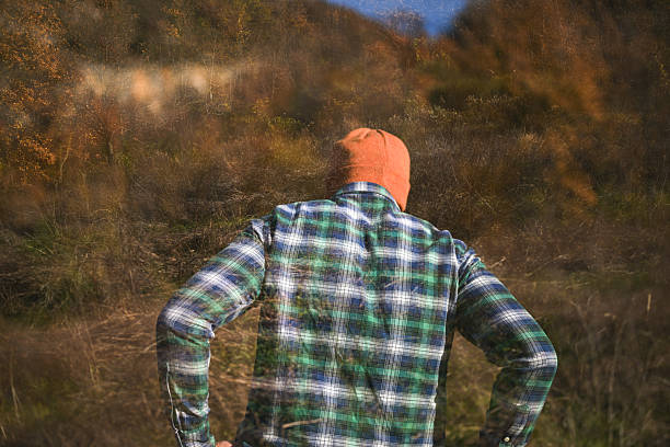 Man standing backwards in a plaid shirt and orange beanie, facing an autumnal forest