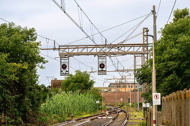Suspended signals show red aspects above curving tracks and overhead wires, as vegetation and equipment line the route near a junction.