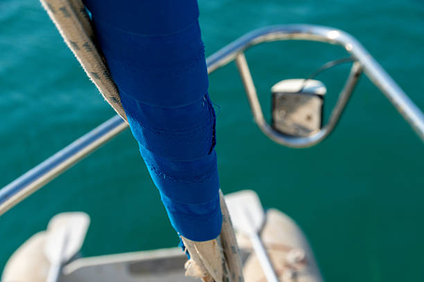Close-up view of a furled sail with damaged seams, attached to stainless steel railing with blue tape, against blurred water background.