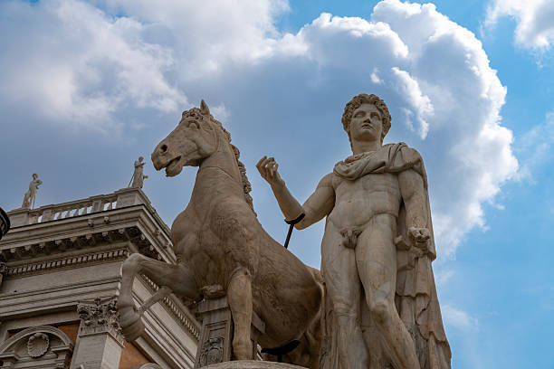 Large marble Statuen of Castor und Pollux with rearing horses rise beside a portico, set against a bright sky with billowing clouds above.