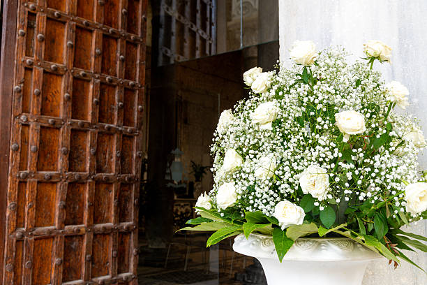 A large bouquet of white roses and tiny white flowers stands in a white urn beside an open wooden church door, revealing a dim interior.