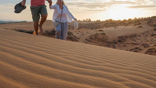 Mature couple hold hands and walk up sand dune at sunset, wave patterns in sand, Red Sand Dunes Park near Mui Ne
