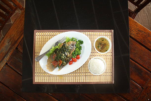 Meal featuring crispy fried fish topped with spicy green sauce, served with white rice and clear soup on a bamboo placemat over a dark table