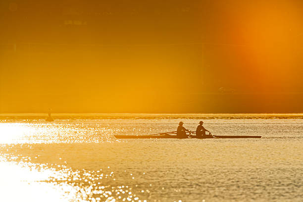 Two women in a double scull rowing on a serene lake, showcasing teamwork and athleticism. The picturesque, captured with a medium shot, backdrop features lush green hills and a quaint church, emphasizing a harmonious blend of nature and sports training.