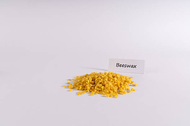 A clean studio shot featuring a pile of pure, golden yellow beeswax pellets on a pristine white background. A white label with 'Beeswax' written in elegant script stands behind the wax, emphasizing its natural origin. The bright, soft lighting highlights the texture of the pastilles, making this image ideal for natural product branding, DIY recipes, cosmetic ingredients, or eco-friendly themes.