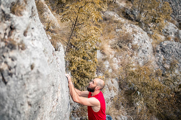Athletic bearded man in a red sleeveless shirt climbing a rocky cliff in nature. Concept of strength, determination, adventure, and outdoor sports