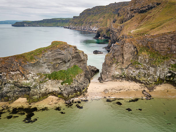 Aerial view of dramatic coastal cliffs and narrow rope bridge spanning turquoise waters near Carrick-a-Rede. Steep rock formations meet the Atlantic Ocean creating a rugged and scenic landscape. Carrick-a-Rede, County Antrim, Northern Ireland, United Kingdom, Western Europe