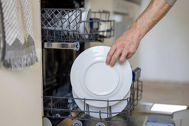 Close-up view of a man's hand placing a white ceramic plate into the rack of an open dishwasher. The scene captures a daily domestic routine in a home kitchen, highlighting themes of housework, cleaning, hygiene, and modern appliance usage.