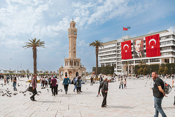 Izmir city center view showing Konak Square crowded with local people and tourists during a national holiday celebration, with the historic Izmir Clock Tower as the main landmark and large Turkish flags and Ataturk portrait displayed on surrounding government buildings, symbolizing Republic Day and national pride, captured in daylight in Izmir, Turkey