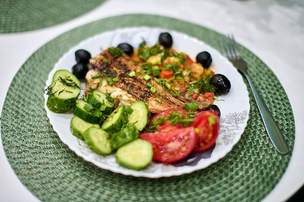 Grilled fish with vibrant vegetables and olives served on white plate. Decorative green mat adds texture. Bright natural light enhances fresh ingredients colors