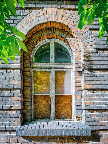 Vintage arched window in an old brick building surrounded by green leaves. Rustic architectural detail in warm sunlight.