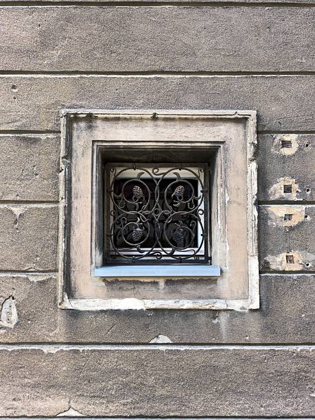 Close-up of a small square window with an ornate wrought iron grill on an aged stone wall. Minimalist architectural detail with texture and patina.