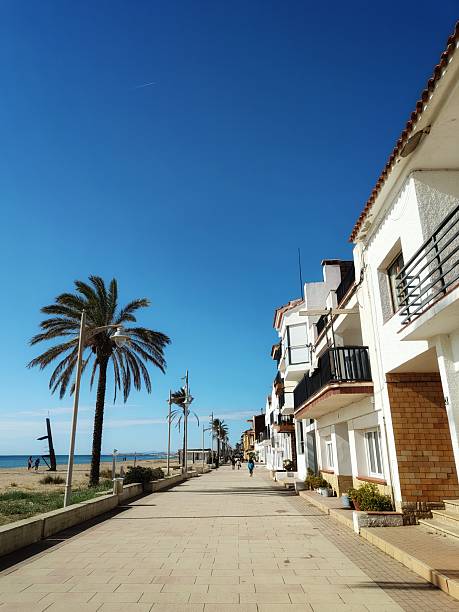 A peaceful view of the seaside promenade in Sant Salvador, Coma-ruga. This shot captures the iconic white Mediterranean architecture, towering palm trees, and the golden beach under a deep blue, cloudless sky.