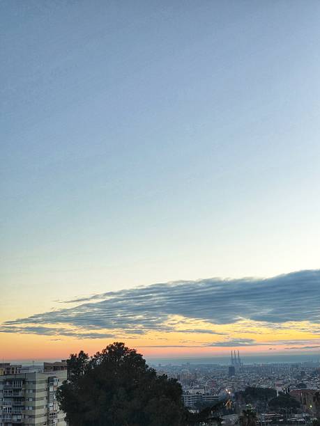 A breathtaking panoramic view of Barcelona at dusk, featuring a soft orange horizon, a unique wave-like cloud formation, and the iconic coastal silhouette transitioning into the twilight.