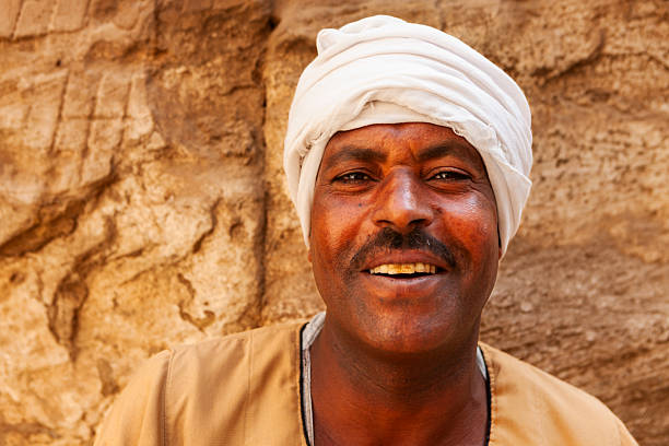 Close-up portrait of a smiling man wearing a white turban and a beige robe, posed against a textured sandstone wall. Warm, golden light brings out the rich tones of his skin and the fine details of his face, conveying a friendly, relaxed expression.