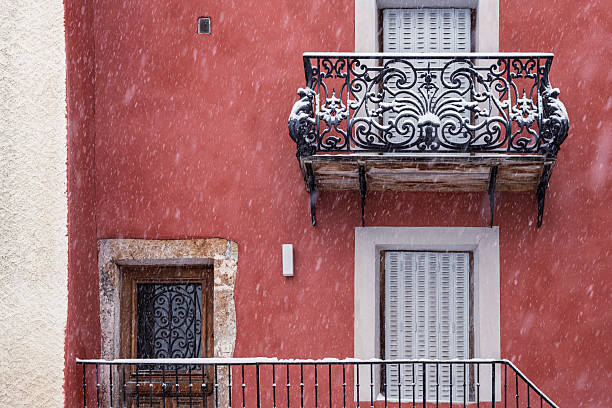 Traditional alpine building facade painted in warm red tones features ornate wrought-iron balcony railing adorned with fresh snowfall in Peisey-Nancroix, France. The classic Savoyard architecture displays characteristic wooden shutters and decorative ironwork, with pristine white snow creating striking contrast against the terracotta-colored wall. The composition captures authentic mountain village character through architectural details and seasonal elements, showcasing the distinctive building styles of French Alpine communities during winter.
