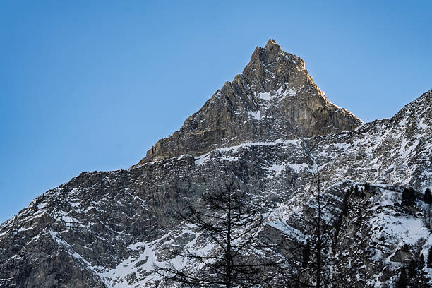 Sharp mountain peak catches golden light on its snow-covered slopes while maintaining its dramatic pyramidal form against deep blue sky in Les Lanches, Peisey-Nancroix, France. The distinctive summit displays perfect snow coverage with subtle terrain features visible across its steep faces, while warm sunlight creates beautiful highlights on the white slopes. The composition captures the peak's impressive geometry and alpine grandeur, with ideal lighting and atmospheric conditions revealing the mountain's magnificent form and the pristine beauty of high-altitude winter scenery.