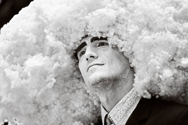 Black and white close-up of male adult performer wearing voluminous cloud - Like headpiece. Expression appears contemplative, emphasizing theatrical and surreal atmosphere