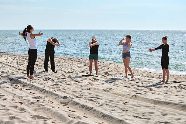 Five adults perform stretching exercises during group yoga session on sunny Baltic Sea beach. Calm sea and clear sky create peaceful outdoor environment