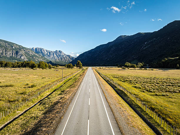 Aerial view of a paved road running between open plains and steep mountain slopes under a clear blue sky. The image highlights the scale and solitude of the Carretera Austral in Chilean Patagonia.