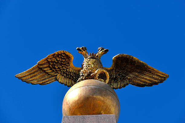 Helsinki, Uusimaa, Finland: the gilded bronze finial atop the Keisarinnankivi (Stone of the Empress) obelisk in the Market Square (Kauppatori). The sculpture depicts the double-headed eagle of the Russian Empire perched on a golden orb. A key historical detail is visible on the eagle's breast: the Coat of Arms of the Grand Duchy of Finland, featuring a lion rampant on a shield. Designed by architect Carl Ludvig Engel and unveiled in 1835, the monument commemorates the 1833 visit of Czarina Alexandra Feodorovna.  The image isolates the golden figures against a clear sky, highlighting the intricate metalwork and imperial symbolism.