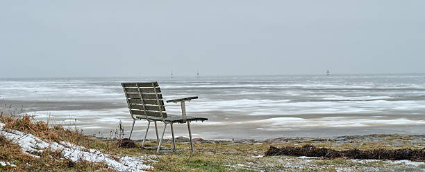 A lone weathered bench sits at the edge of a cold coast, with patches of snow and grass in the foreground and a calm, icy sea stretching to the horizon. The scene conveys solitude, calm, and reflection in a Nordic seaside setting, perfect for themes of winter, silence, and nature.