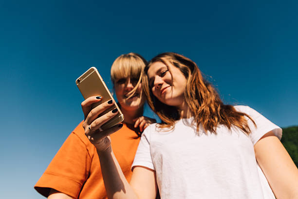 Two young adult women smiling and holding a smartphone, capturing a selfie or recording content on social media with a clear blue sky in the background