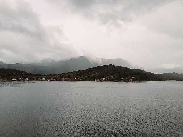Classic red and white Norwegian cabins and boathouses sit at the edge of the water with steep, cloud-covered mountains in the background.