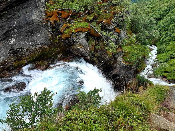 A high-angle perspective looking down a rushing waterfall as it plunges over mossy rocks and cliffs into a green valley below.