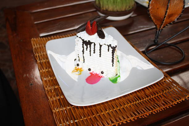 Sweet dessert cake slice, frosted with white cream and chocolate drizzle, decorated with a whimsical duck face Served on a white plate over a woven placemat