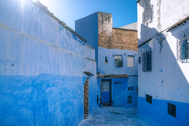 A sunlit, blue-washed alley in Chefchaouen, Morocco. Narrow street with blue walls, windows with iron grilles, and a doorway, capturing tranquil, photogenic Moroccan architecture and travel allure.
