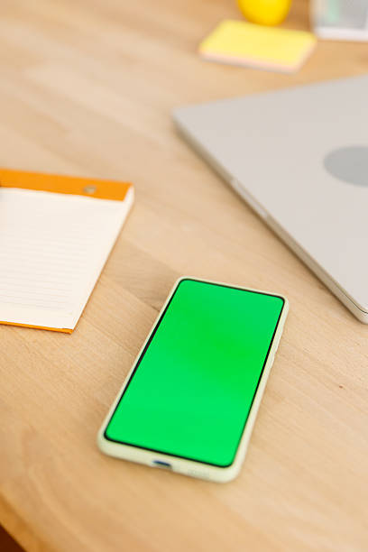 Smartphone displaying a green screen for custom content placed on a wooden desk next to a closed laptop and a notebook, creating a modern workspace setup