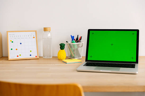 Laptop with green screen for copy space on a clean wooden desk with monthly planner, water bottle and stationery, minimalist home office setup for remote work and planning
