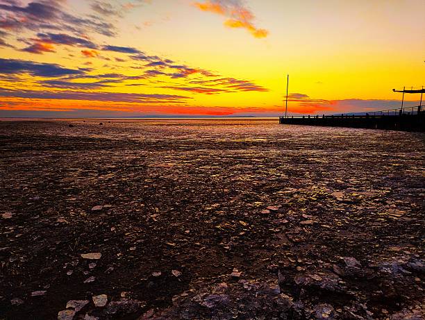 Wide view of Neusiedler See frozen into overlapping plates of broken ice glowing in orange sunset light, with long pier and pastel clouds on the horizon at Seegelhafen West in winter.