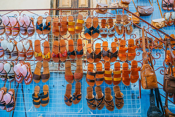 Colorful collection of leather sandals neatly arranged on a metal grid display in a vibrant Moroccan market. Warm browns and bright straps contrast against blue walls, capturing casual summer fashion and handmade craftsmanship.