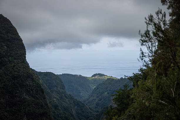 Sunlit hilltop village on Madeira island above deep green valley, low storm clouds hang over the Atlantic horizon with forested cliffs framing distant houses