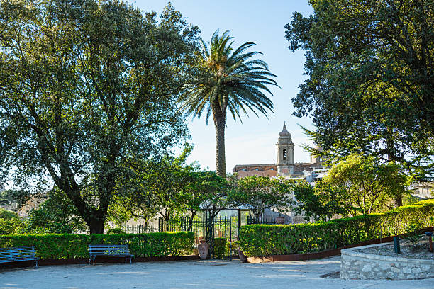 A tranquil Sicilian park scene featuring lush trees, a tall palm, and a distant historic church bell tower. Benches and hedges provide a peaceful setting with a glimpse of old town architecture.