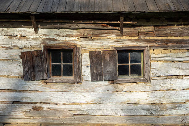 Exterior wall of a traditional wooden house built with horizontal timber beams and clay infill. Two small wooden windows with rustic shutters highlight old rural architecture and handcrafted construction. Weathered wood texture and historic building details.