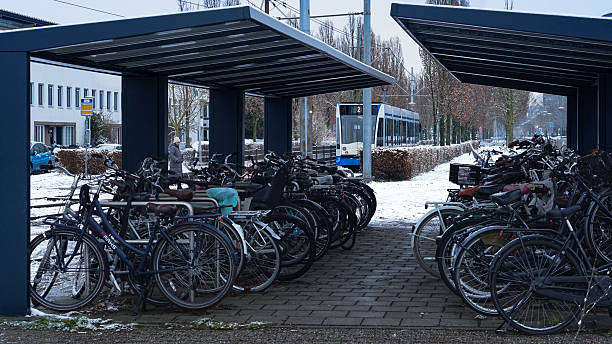 A covered bicycle parking area is seen at the terminus of tram line 2 in the New Sloten district of Amsterdam, Netherlands, on January 29, 2026. Numerous bicycles are parked under two shelter structures, with snow covering the ground and creating a winter scene. Residential buildings and leafless trees are visible in the background, indicating cold seasonal weather. The image documents everyday urban transportation infrastructure and local commuting conditions during snowfall