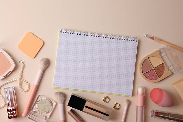 Top View Cosmetics Layout With Brushes, Palettes, Sponge, Lipstick And Notebook Arranged On Soft Beige Surface