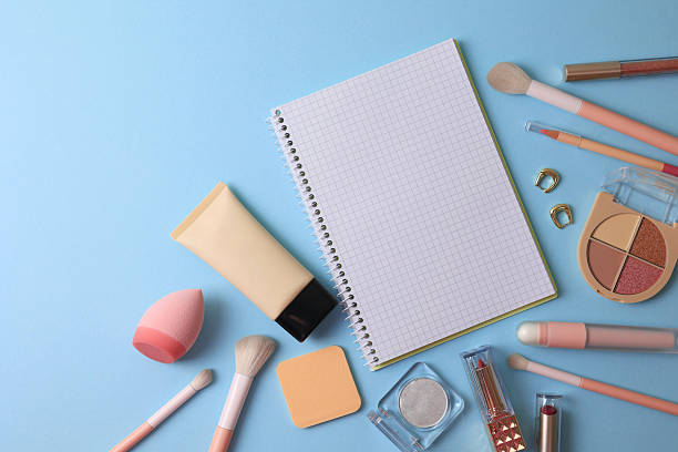 Top View Cosmetics Layout With Brushes, Palettes, Sponge, Lipstick And Notebook Arranged On Soft Beige Surface