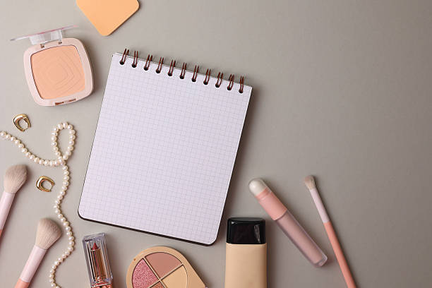 Top View Cosmetics Layout With Brushes, Palettes, Sponge, Lipstick And Notebook Arranged On Soft Beige Surface