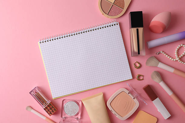 Top View Cosmetics Layout With Brushes, Palettes, Sponge, Lipstick And Notebook Arranged On Soft Beige Surface
