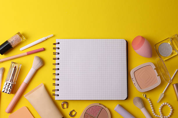 Top View Cosmetics Layout With Brushes, Palettes, Sponge, Lipstick And Notebook Arranged On Soft Beige Surface