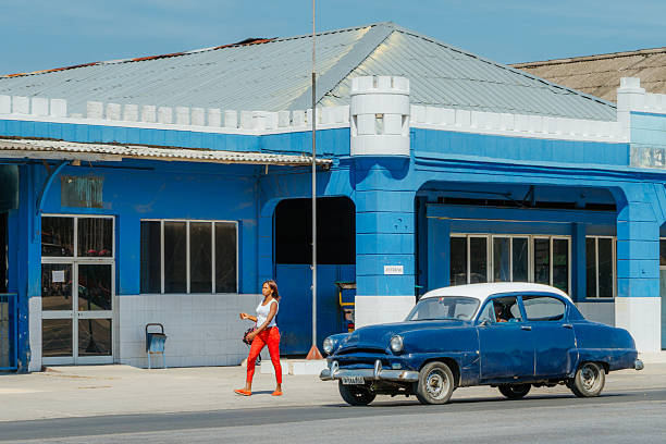 Santiago de Cuba, Cuba on February 15, 2017: A bright blue vintage car drives along a sunlit Cuban street while a woman in red pants walks past a blue building. The scene captures everyday life, color, and retro charm in Santiago de Cuba.