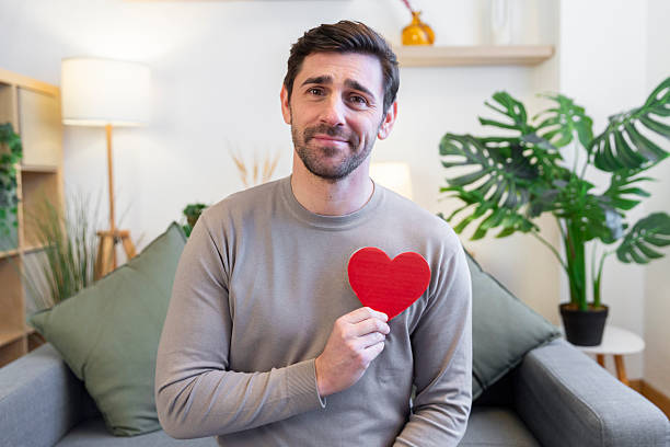 Man holding a red heart to his chest in a living room, smiling at camera
