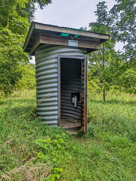 A corrugated metal outhouse made from an old culvert sits in a grassy clearing, framed by leafy trees. Rustic outdoor restroom in a rural setting emphasizing simplicity, solitude and natural surroundings.